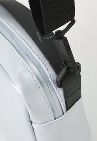 Sac gris, brillant, avec des accents noirs et une sangle texturée. Présente une finition lisse et des coutures détaillées le long du bord. Boucle ajustable.