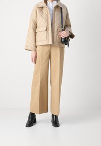 Beige gewatteerde jas met drukknoopsluiting en zijzak, gecombineerd met lichtbruine wijde broek en zwarte enkellaarsjes met hak.