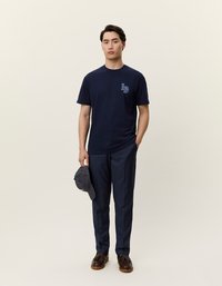 Jūras zils kokvilnas T-krekls ar gaiši zilu logo, kombinēts ar tumšiem biksēm un brūniem apaviem. Roktā pa kreisi turētā strīpainā cepure.