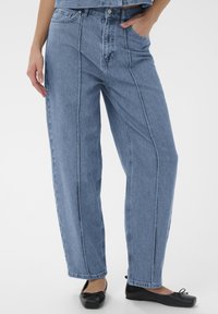 Frau in hochgeschnittenen, lockeren blauen Jeans mit Vorderfalten, die die Hände in den Taschen hat, kombiniert mit schwarzen Ballerinas vor einem weißen Hintergrund.