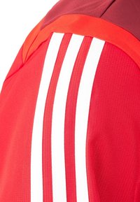 Rote Sportjacke mit drei weißen Streifen auf der Schulter. Strukturiertes Gewebe mit glatter Oberfläche und kontrastierenden roten Akzenten.
