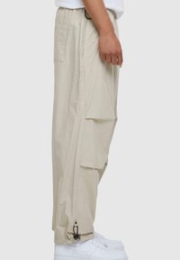 Beige cargo bukser med store sidelommer, elastisk talje og justerbare manchetter. Båret med hvide sneakers. Glat, let stof.