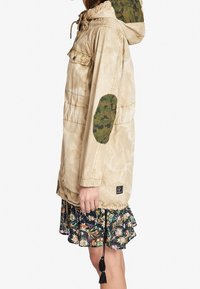Beżowa kurtka z kapturem, ze sznurkiem, bocznymi kieszeniami i zielonymi łatami w kamuflażu na łokciach, zestawiona z kwiecistą spódnicą z falbanami. Tkanina o fakturze.