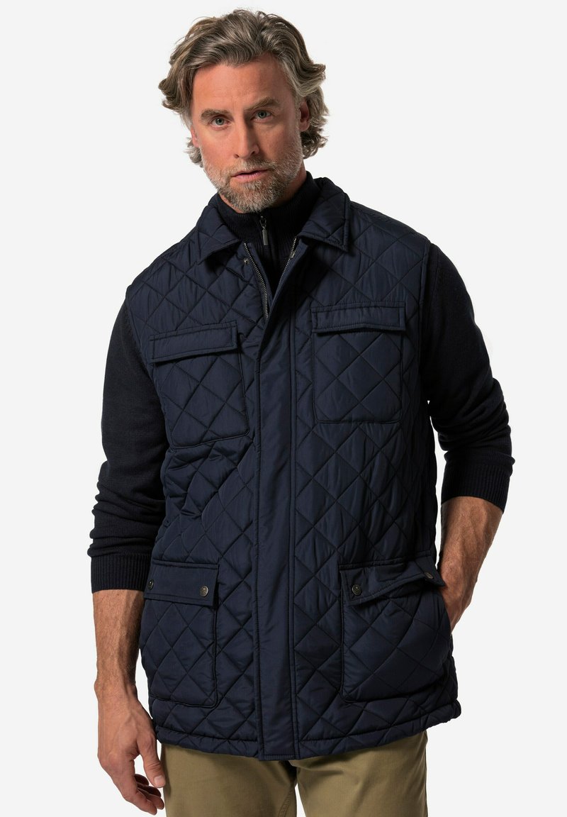 Marinblå quiltad väst med två framfickor, krage och dragkedjestängning. Texturerad tyg med diamantskärm, buren över en svart tröja.