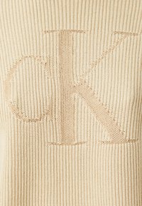 Maglione lavorato a maglia color crema con texture a coste, caratterizzato da un grande logo "CK" beige, leggermente in rilievo. Tessuto morbido e leggero.
