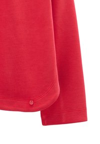 Czerwony długi rękaw koszuli z zaokrąglonym dołem i małą okrągłą czerwoną metką z logo blisko dolnej krawędzi na białym tle.