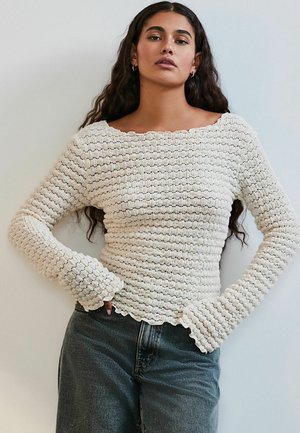 Langarm, crèmekleurig gebreid topje met een gestructureerd patroon, golvende halslijn en zoom. Gecombineerd met hoge taille spijkerjeans.