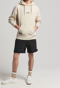 Beige bavlněná mikina s kapucí a logem "SUPERDRY", vpředu klokaní kapsa, kombinovaná s černými kraťasy a bílými teniskami, doplněná vícebarevnými pruhovanými ponožkami.
