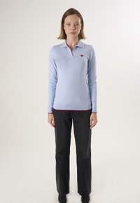 Polo shirt azzurro chiaro a maniche lunghe con colletto, con piccolo emblema a forma di cuore rosso sul petto sinistro e rifiniture rosse all'orlo. Abbinato a jeans neri.