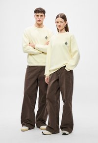 Poros modelių, vilkinčių derančius šviesiai geltonos spalvos džemperius su juodu deimanto logotipu, rudus plačių kojų kelnes ir smėlio spalvos sportbačius.