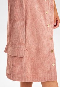 Korddräkt i mjuk rosa med vertikala ribbor, med guldtonade knappar och en sidoflappar.