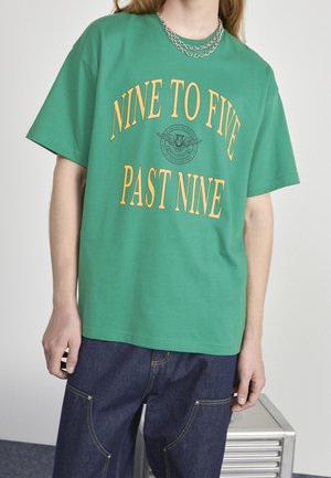Personne portant un t-shirt vert avec un texte jaune « NINE TO FIVE PAST NINE » et un jean bleu, debout à côté d'une petite table en métal.