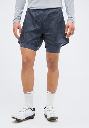 Férfi, fekete réteges rövidnadrágot, hosszú ujjú szürke inget, fehér zoknit és fehér kerékpáros cipőt visel, sima háttér előtt áll.