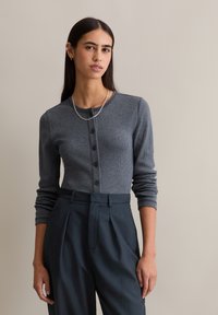 Szary prążkowany kardigan z długimi rękawami, zapinany na guziki z okrągłym dekoltem, zestawiony z ciemnymi, spodniami z wysokim stanem. Dodatkiem jest srebrny łańcuszek.