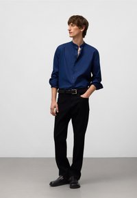Donkerblauw overhemd met knopen en een staande kraag, zwarte broek en zwarte instappers. Glad materiaal, slim fit en opgerolde mouwen.