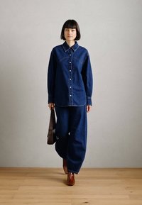 Τζιν πουκάμισο και παντελόνι με φαρδιά πόδια σε σκούρο μπλε. Το πουκάμισο διαθέτει γιακά και μπροστινά κουμπιά, ενώ το παντελόνι έχει χαλαρή εφαρμογή. Καφέ τσάντα και παπούτσια.