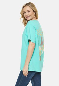 Turkos t-shirt med kort ärm och en lekfull grafisk design på baksidan i orange och beige nyanser, med avslappnad passform och mjuk bomullstextur.