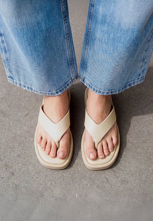 Pies descalzos en sandalias color crema con tiras anchas, llevados con vaqueros de mezclilla azul claro, de pie sobre un suelo de hormigón gris.