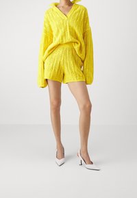 Gul stickad tröja och shorts-set med ett strukturerat mönster. Har en V-ringning och långa ärmar, ihop med vita spetsiga klackar.