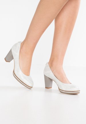 Jalat yang mengenakan sepatu blok tumit tertutup berwarna abu-abu muda dengan tekstur, satu kaki sedikit diangkat di latar belakang putih polos.