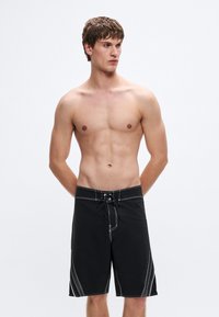 Μαύρα σορτς με λευκή ραφή, με σούρα στη μέση, πλαϊνές τσέπες και χαλαρή εφαρμογή μέχρι το γόνατο. Απλό γεωμετρικό σχέδιο.