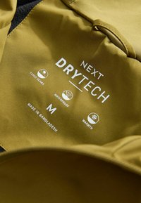 Eticheta din material verde măsliniu prezintă brandul "Next Drytech", simboluri pentru impermeabilitate, căldură, cusături lipite și mărimea "M", cu inscripția "Fabricat în Bangladesh."