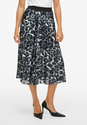Vrouw die een geplooide midi-rok draagt met een zwarte en grijze luipaardprint, gecombineerd met zwarte puntige hoge hakken.