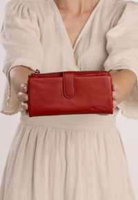 Cartera de cuero rojo con una textura suave, cierre de cremallera y una solapa frontal. Presenta un sutil logo en la esquina inferior derecha.