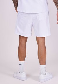 Pantalones cortos blancos hechos de un tejido ligero, con un diseño sencillo. Tienen un bolsillo trasero y el modelo lleva zapatillas blancas con detalles negros.