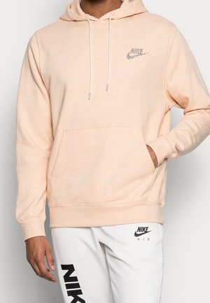 Mężczyzna w beżowej bluzie z kapturem Nike z przednią kieszenią oraz białych spodniach Nike Air z czarnym logo na udzie na jednolitym tle.