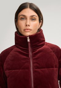 Giacca imbottita bordeaux con colletto alto e zip, realizzata in tessuto morbido e testurizzato. Caratterizzata da un design trapuntato e da una silhouette elegante.