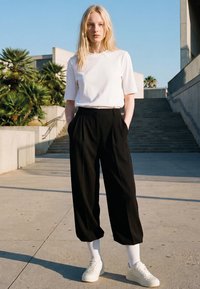 Woman with long blonde hair wearing a white T-shirt, black wide-leg pants, white socks, and white sneakers stands outdoors near stairs.