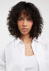 Camisa blanca de lino con botones en el delantero, mangas cortas y una textura sutil, llevada sobre una camiseta de tirantes blanca de canalé. Cabello rizado natural enmarcando el rostro.