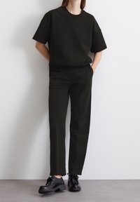 Černý bavlněný oversized top s krátkými rukávy, kombinovaný s černými kalhotami na míru a černými šněrovacími botami, představující minimalistický design.