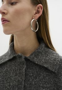 Maglione grigio con texture, colletto e bottoni neri; modello che indossa grandi orecchini a cerchio d'argento. Capelli lisci, sfondo neutro.