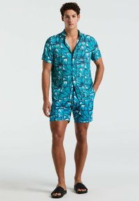 Turquoise set bestaande uit een shirt met korte mouwen en shorts, met een gedurfde bladdessin, gemaakt van lichtgewicht stof. Gecombineerd met zwarte instapsandalen.