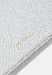 Hvit teksturert skinnpose med et gullforgylt "Coccinelle"-logo, med glatte kanter og høy kvalitet i utførelsen.