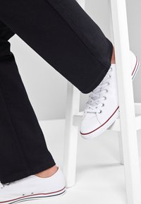 Weiße Leinensneaker mit runder Zehenpartie, weißen Schnürsenkeln und roten Akzenten, kombiniert mit schwarzen Hosen, präsentiert auf einem weißen Hocker.