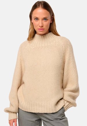 Un pull en tricot crème épais avec un col haut en côtes et des épaules tombantes, présentant un motif texturé et des manches larges.