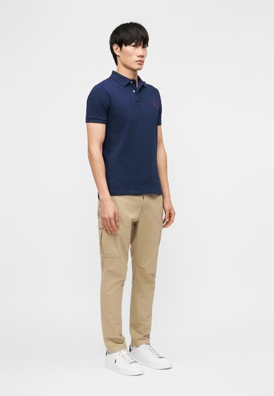 Uomo in piedi, indossa una polo blu navy, pantaloni cargo beige e sneakers bianche, rivolto leggermente verso sinistra su uno sfondo bianco uniforme.