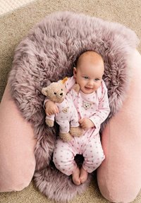 Pink baby onesie with star pattern, cuddling a teddy bear. Seated on a fluffy, light pink cushion with a textured, full background.