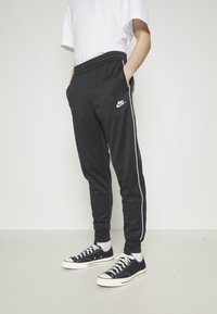 Osoba v černých trenýrkách Nike s bílými bočními pruhy, bílými ponožkami, černobílých teniskách a jednoduchém bílém tričku, stojící uvnitř.
