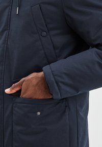 Giacca isolante blu navy con chiusura a zip, tasca laterale e dettaglio con bottone. Tessuto liscio e design strutturato con una vestibilità comoda.