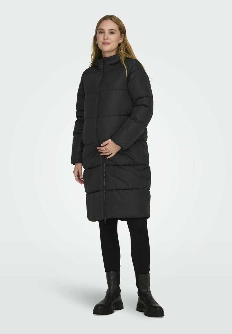 Abrigo largo acolchado negro con cuello alto, cierre de cremallera en el frente y costuras horizontales. Llevado sobre leggings negros y botas gruesas negras.