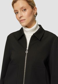 Vrouw met blond haar draagt zilveren creolen, een witte coltrui en een zwarte jas met een ritssluiting aan de voorkant en grote zakken.