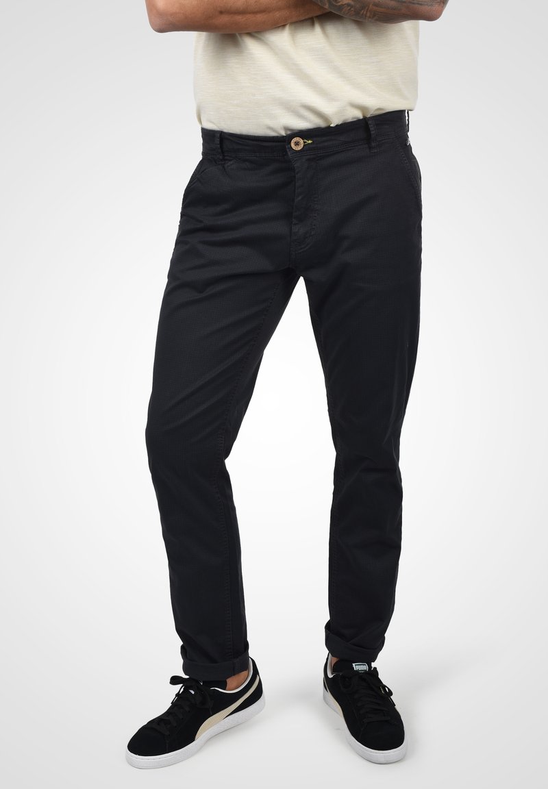 Pantalon en mélange de coton noir à coupe droite, avec des poches avant et une fermeture à bouton unique. Porté avec des baskets noires et blanches.