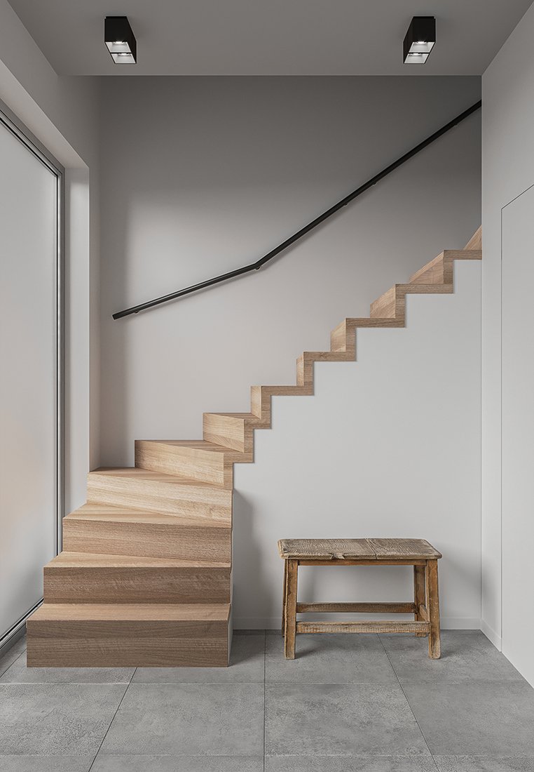 Escalera de madera con peldaños angulares, barandilla negra y suelo de azulejos grises. Un taburete de madera rústico está colocado en la base de las escaleras.