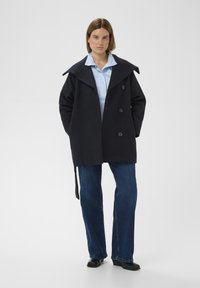 Tmavomodrý kabát zo zmesi vlny s veľkým golierom, dvojradovým zapínaním na gombíky a voľným strihom; nosený cez svetlomodrú košeľu s golierom a džínsy.