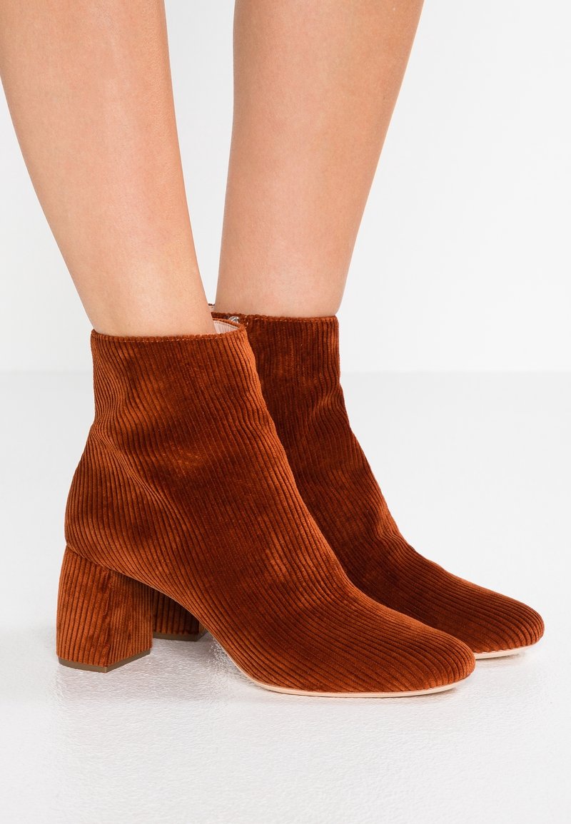 Bottines en velours côtelé marron à talons blocs portées sur jambes nues sur fond blanc.