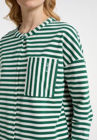 Maglietta a righe verdi e bianche con scollatura rotonda, bottoni frontali, una tasca sul petto e logo dell'ancora sulla tasca. Tessuto morbido.